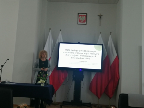 Anna Gawryluk – pani Anna Gawryluk przy stole konferencyjnym na tle biało-czerwonych flag, obok monitor z tematem wykładu.