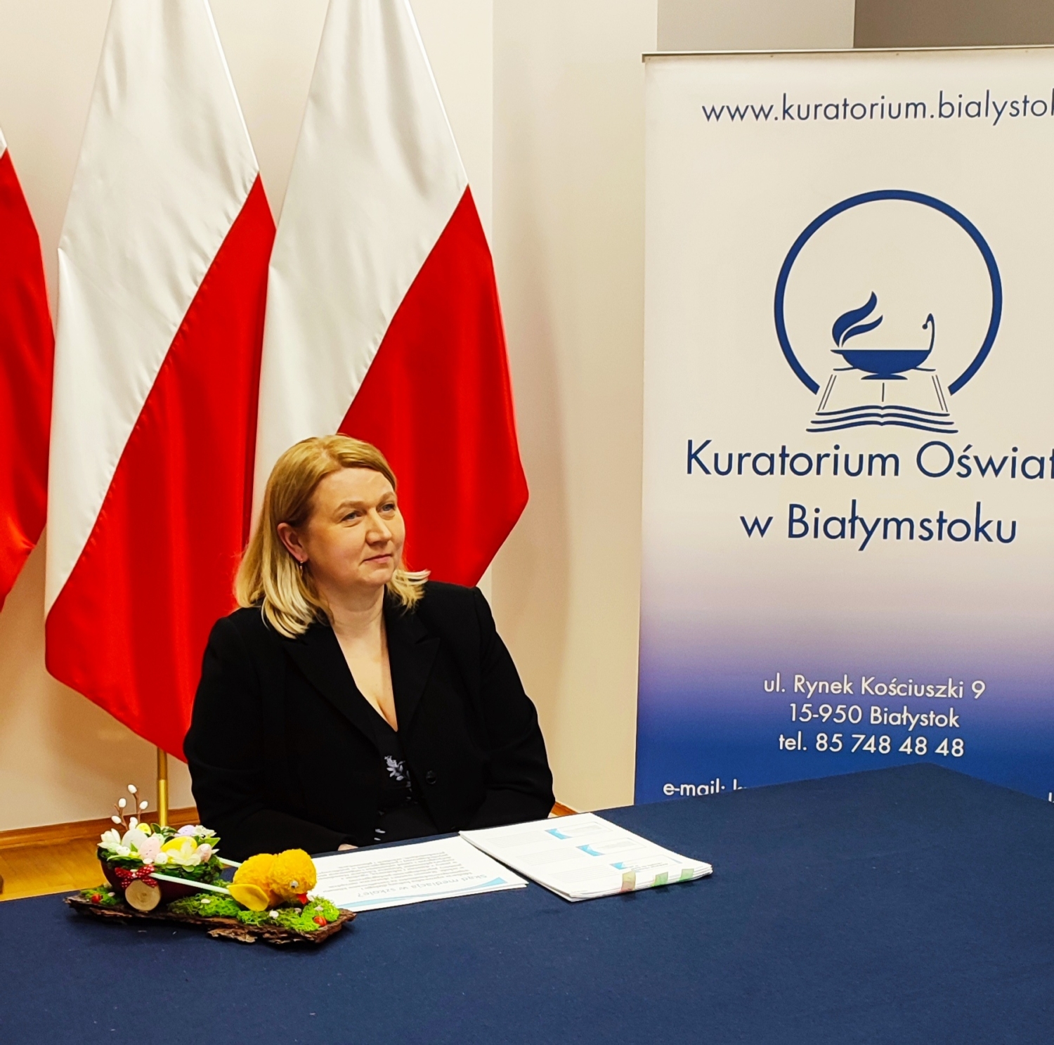 Pani Milena Kucharczyk – mediator sądowy siedzi przy stole na tle flag biało-czerwonych, z boku ustawiony baner z logo Kuratorium Oświaty w Białymstoku.