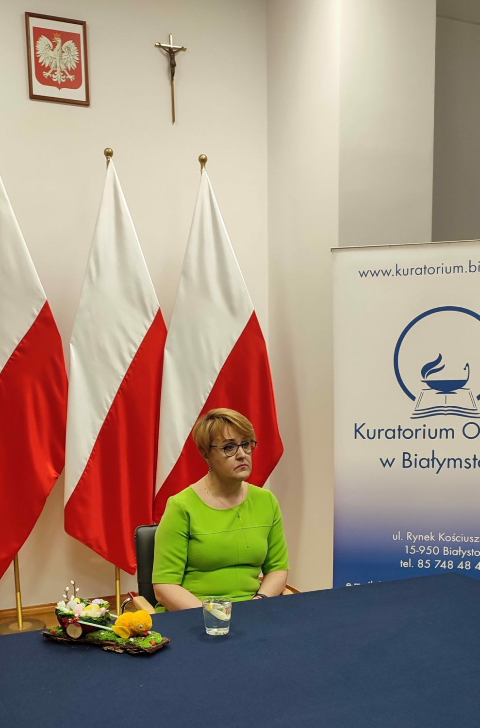 Pani psycholog Katarzyna Kalinowska siedzi przy stole, na tle flag biało-czerwonych, z boku ustawiony baner z logo Kuratorium Oświaty w Białymstoku.